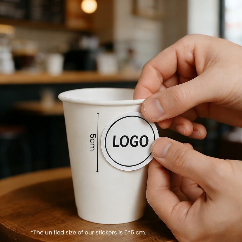 Applying a logo sticker to a 10oz single wall paper cup on a wooden table inside a coffee shop.