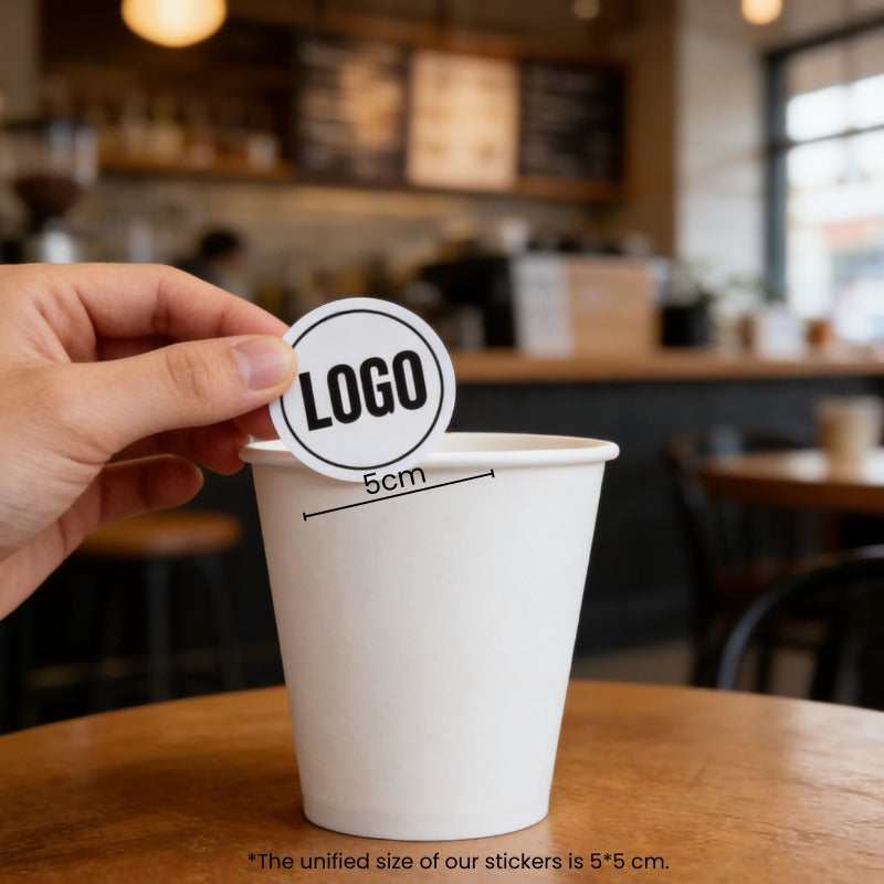 Hand applying a 2-inch logo sticker onto an 8oz single wall white paper cup in a café setting.
