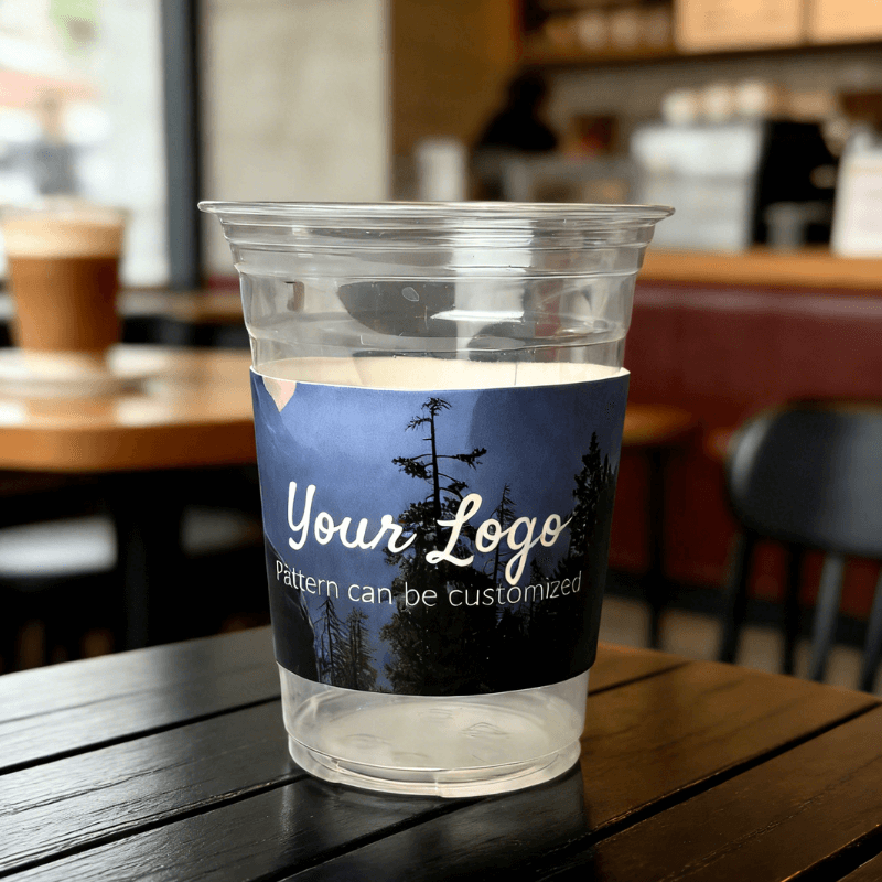 Clear plastic cup on a cafe table wrapped with a night-forest cup sleeve featuring a customizable logo.