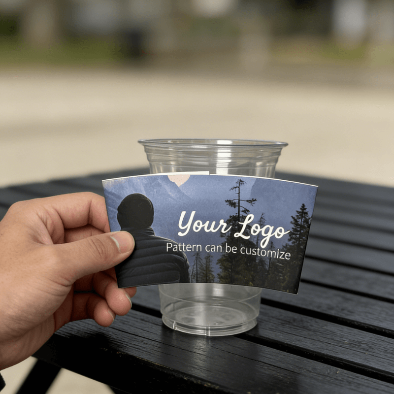 Hand holding a custom logo night-forest cup sleeve in front of a clear plastic cup outdoors.