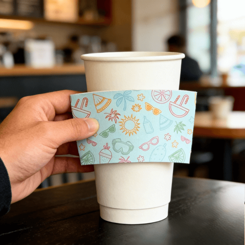 Hand holding a light-blue summer icon cup sleeve in front of a white paper cup on a cafe table.