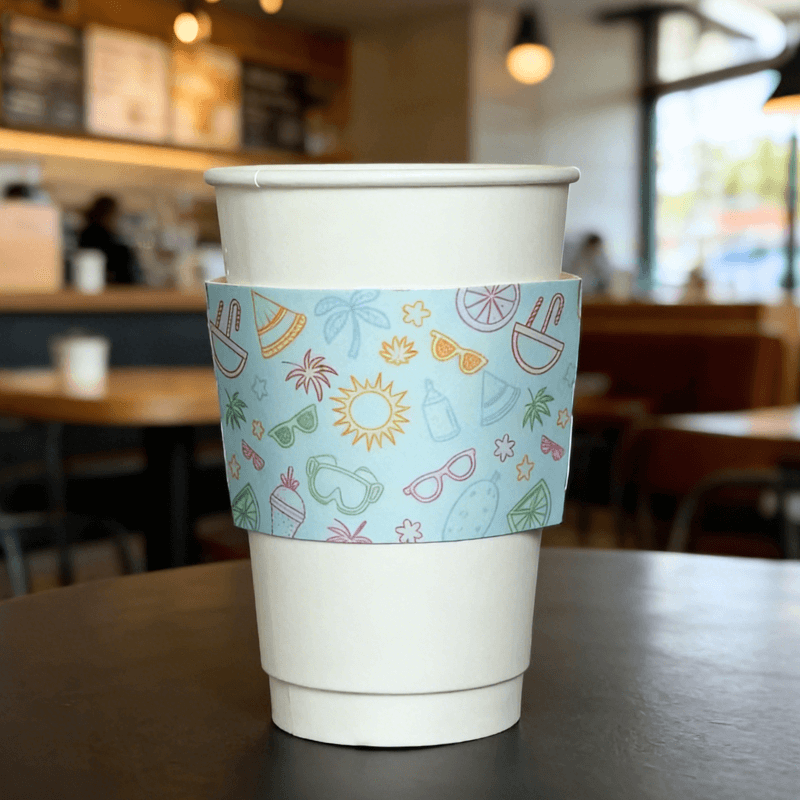 White paper cup wrapped with a light-blue summer icon cup sleeve in a cafe setting.