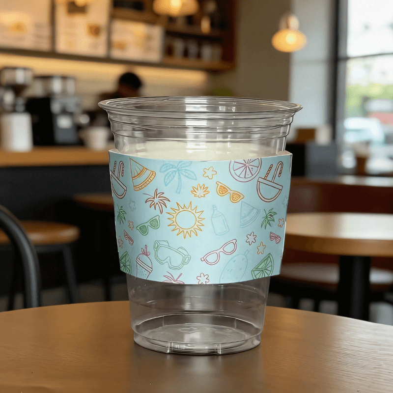 Clear plastic cup wrapped with a light-blue summer icon cup sleeve on a cafe table.