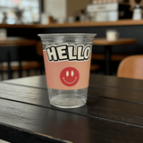 Clear plastic cup on a café table with a pink sleeve that says “HELLO” and a red smiley face; background is softly blurred.