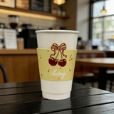 White paper cup on a café table with a yellow sleeve featuring a bow and cherries and the word “cherry”; background is softly blurred.