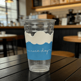 Clear plastic cup on a café table with a blue sleeve topped with a white cloud shape and the phrase “a nice day”; background is softly blurred.