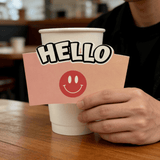 White paper cup on a café table with a pink sleeve that says “HELLO” and a red smiley face; background is softly blurred.