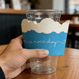 Hand holding a clear plastic cup with a blue sleeve topped with a white cloud shape and the phrase “a nice day.