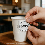 Applying a logo sticker to a 10oz single wall paper cup on a wooden table inside a coffee shop.
