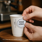 12oz double wall paper cup with a centered logo sticker shown in a cozy coffee shop environment.