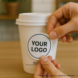 Close-up of hands placing a round logo sticker onto a 12oz single wall paper cup in a café scene.