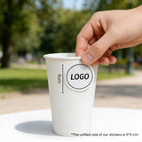 Applying a logo sticker to a large 16oz single wall white paper cup in a café setting.
