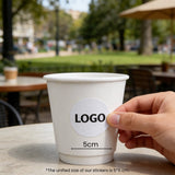 Hand applying a 2-inch logo sticker onto an 8oz double wall white paper cup in a café setting.
