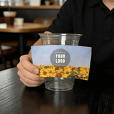 Hand holding a sunflower-field cup sleeve with a customizable logo in front of a clear plastic cup in a cafe.
