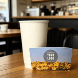 Sunflower-field cup sleeve with a customizable logo displayed in front of a plain white paper cup on a wooden table.