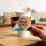 Hand holding a kraft cup sleeve with colorful abstract shapes and a customizable logo in front of a white paper cup.