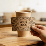 Hand holding a vintage floral-pattern cup sleeve with a customizable logo in front of a kraft paper cup on a wooden table.