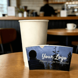 Custom logo forest-design cup sleeve shown in front of a plain white paper cup on a wooden table.