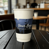 White paper cup wrapped with a dark night-forest cup sleeve showing a customizable logo in a cafe setting.