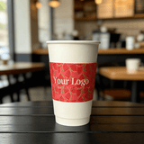 White paper cup on a cafe table wrapped with a red watermelon-pattern cup sleeve featuring a customizable logo