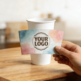 Hand holding a customizable logo cup sleeve with a watercolor gradient design in front of a white paper cup.