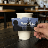 Hand holding a custom logo night-forest cup sleeve in front of a white paper cup in a cafe.