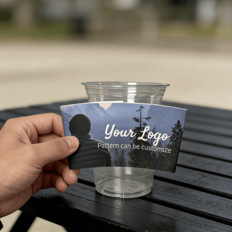 Hand holding a custom logo night-forest cup sleeve in front of a clear plastic cup outdoors.