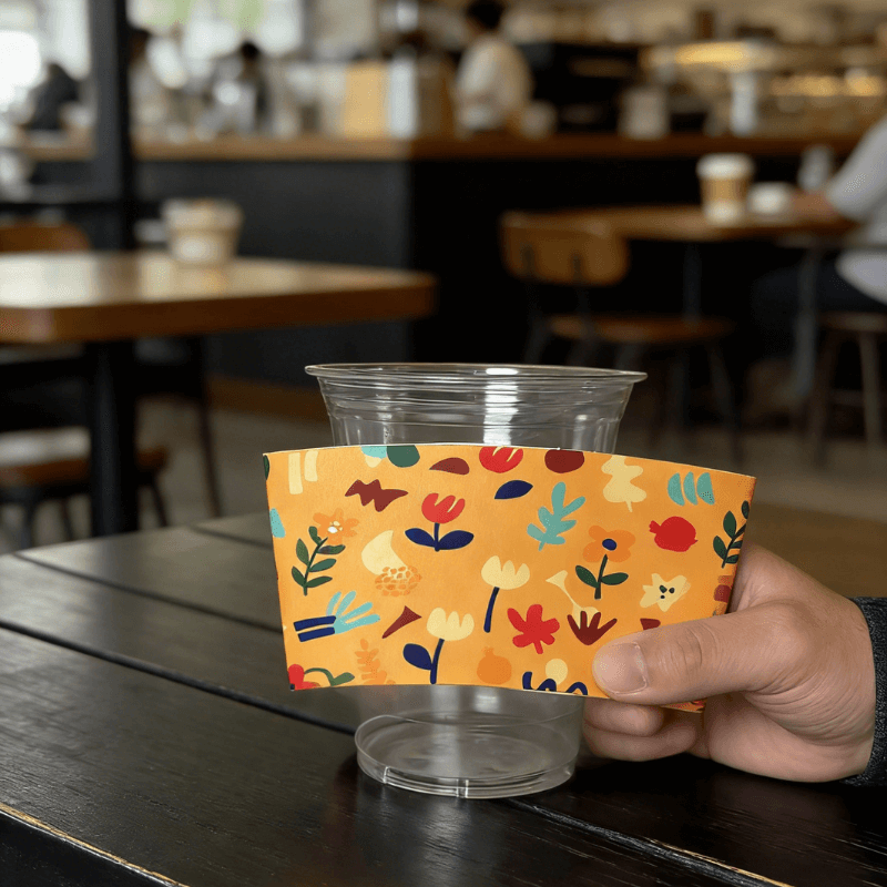 Hand holding an orange floral-pattern cup sleeve against a clear plastic cup in a cafe.