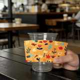 Hand holding an orange floral-pattern cup sleeve against a clear plastic cup in a cafe.