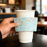 Hand holding a light-blue summer icon cup sleeve in front of a white paper cup on a cafe table.