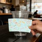 Hand holding a light-blue summer icon cup sleeve in front of a clear plastic cup on a cafe table.