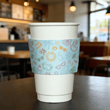 White paper cup wrapped with a light-blue summer icon cup sleeve in a cafe setting.