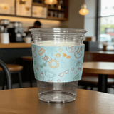 Clear plastic cup wrapped with a light-blue summer icon cup sleeve on a cafe table.