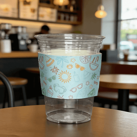 Clear plastic cup wrapped with a light-blue summer icon cup sleeve on a cafe table.